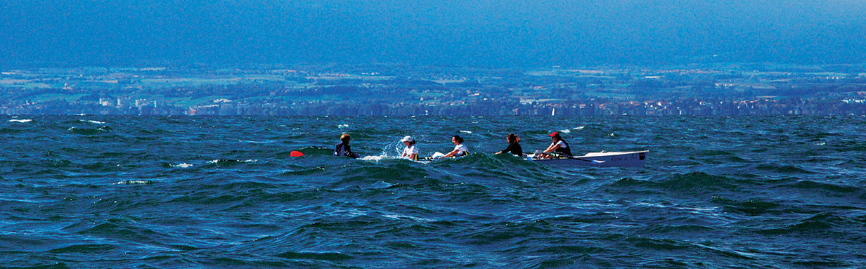 Five rowers in a rowing boat on choppy dark blue open water with a distant coastline visible on the horizon