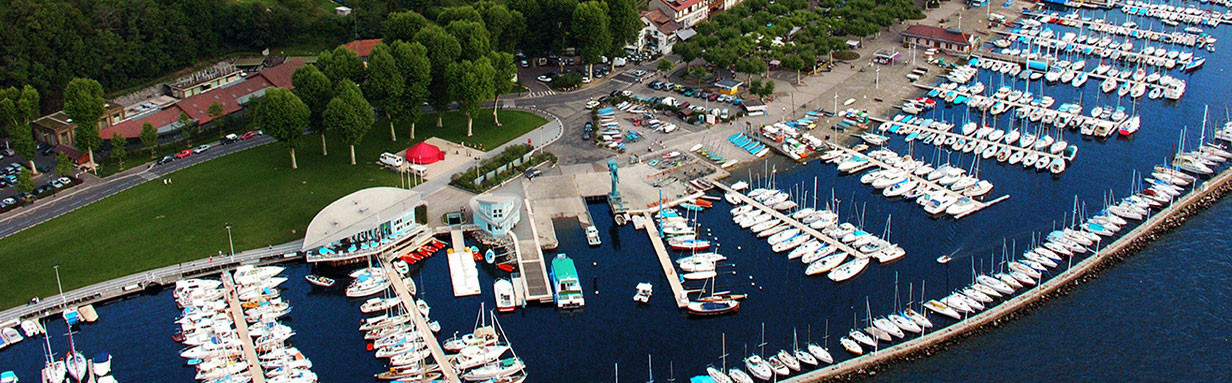 Aerial view of a marina packed with white sailboats and motorboats along a lakeside, green park area and buildings nearby