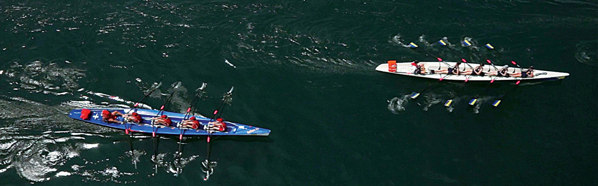 Two rowing crews in narrow boats, one blue and one white, cutting across dark green lake water under soft golden-hour light