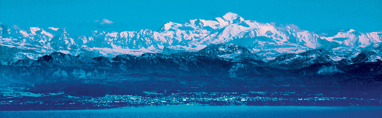 Snow-capped mountain range under a blue sky with a lakeside town below, cool blue toned landscape